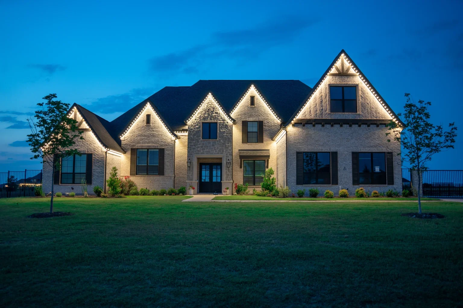 A two-story home at dusk with warm white permanent lights along every roofline and gable